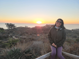 Photo of Paulette Morales at sunset sitting on a fence with Torrey Pines State Park in the background. 